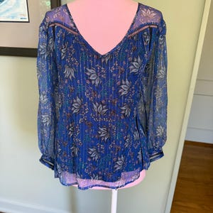 May include: A blue floral blouse with a V-neck and long sleeves. The sheer fabric features a pattern of leaves and vertical stripes. The sleeves and neckline are trimmed with a delicate, patterned border. The blouse is displayed on a pink mannequin.