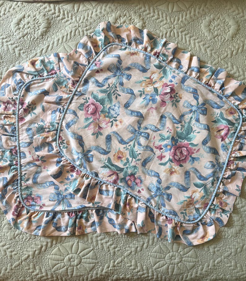 May include: Two pillowcases with a floral and ribbon pattern. The pillowcases have a light peach background with blue ribbons and floral bouquets in pink, blue, and yellow. The edges are ruffled and trimmed with a blue piping.