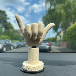 May include: A beige-colored, 3D-printed hand in the shaka gesture, mounted on a stand. The base of the stand reads "HANG LOOSE" in capital letters. The object is designed to be placed on a car dashboard.