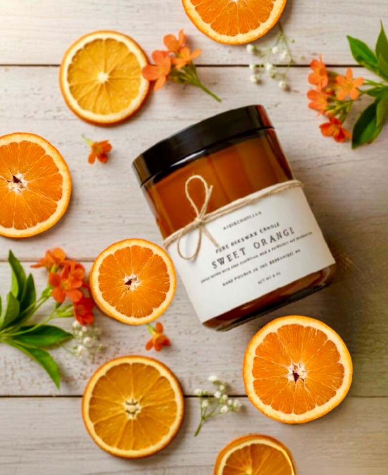 May include: A cylindrical amber glass candle jar with a black lid, wrapped in a white paper label tied with twine. The label reads Sweet Orange, Pure Beeswax Candle, and Net Wt 8 oz. The candle is surrounded by several bright orange slices.