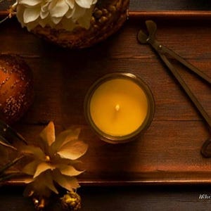 May include: A yellow candle in a glass jar sits on a dark wooden tray alongside a pair of metal wick trimmers, a textured bronze-colored sphere, and two decorative flowers with light-colored petals arranged in a warm, dimly lit setting.