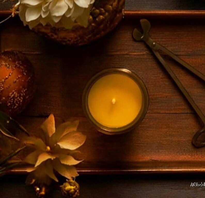 May include: A yellow candle in a glass jar sits on a dark wooden tray alongside a pair of metal wick trimmers, a textured bronze-colored sphere, and two decorative flowers with light-colored petals arranged in a warm, dimly lit setting.