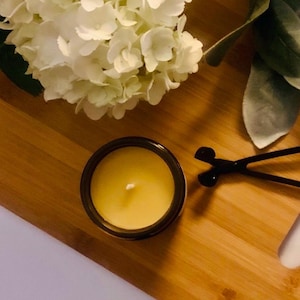 May include: A yellow candle in a dark brown glass jar with a single white wick, positioned next to a black metal wick trimmer. The candle is surrounded by white hydrangea flowers and green leaves, creating a bright and natural composition.