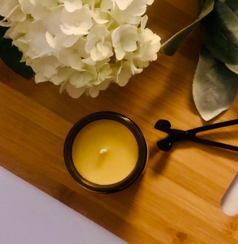 May include: A yellow candle in a dark brown glass jar with a single white wick, positioned next to a black metal wick trimmer. The candle is surrounded by white hydrangea flowers and green leaves, creating a bright and natural composition.