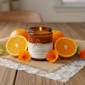May include: A lit candle in a brown glass jar with a white label reading Sweet Orange, surrounded by two halved oranges, one whole orange, and two small orange flowers. The candle features a simple twine bow tied around the jar's neck.
