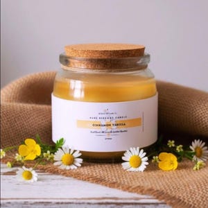 May include: A clear glass jar candle filled with yellow beeswax, topped with a round cork lid. The white label features the text Cinnamon Vanilla and a small flower graphic. The candle is surrounded by small white and yellow daisy-like flowers.