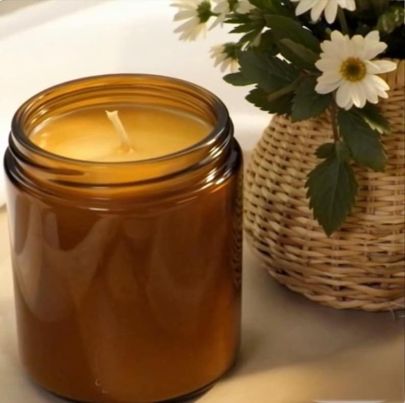 May include: A cylindrical amber glass candle jar filled with smooth yellow wax and a single centered cotton wick. The jar is positioned next to a woven light brown wicker basket containing a bouquet of white daisies with yellow centers.