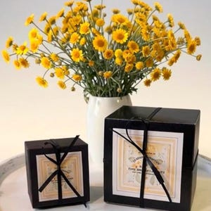 May include: Two black square gift boxes tied with thin black ribbon, each featuring a square label with light yellow borders and printed text. Behind the boxes sits a white vase filled with a large bouquet of bright yellow daisy-like flowers.