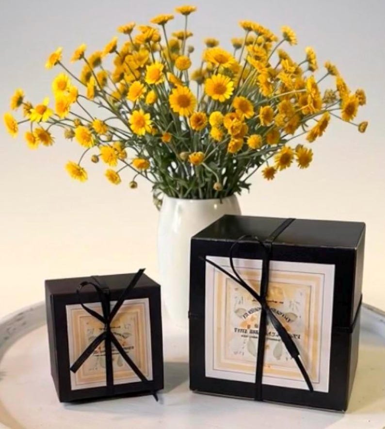 May include: Two black square gift boxes tied with thin black ribbon, each featuring a square label with light yellow borders and printed text. Behind the boxes sits a white vase filled with a large bouquet of bright yellow daisy-like flowers.