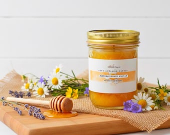 May include: A glass jar candle with a gold lid and a white label reading Natural Honey Nectar. The candle contains yellow wax and sits next to a wooden honey dipper with a drop of honey, surrounded by white daisies and purple lavender flowers.