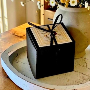 May include: A square black gift box with a black ribbon tied in a bow on top. The box features a square label with printed text and a floral design. The box is positioned on a white circular tray next to a ceramic vase filled with yellow and white flowers.