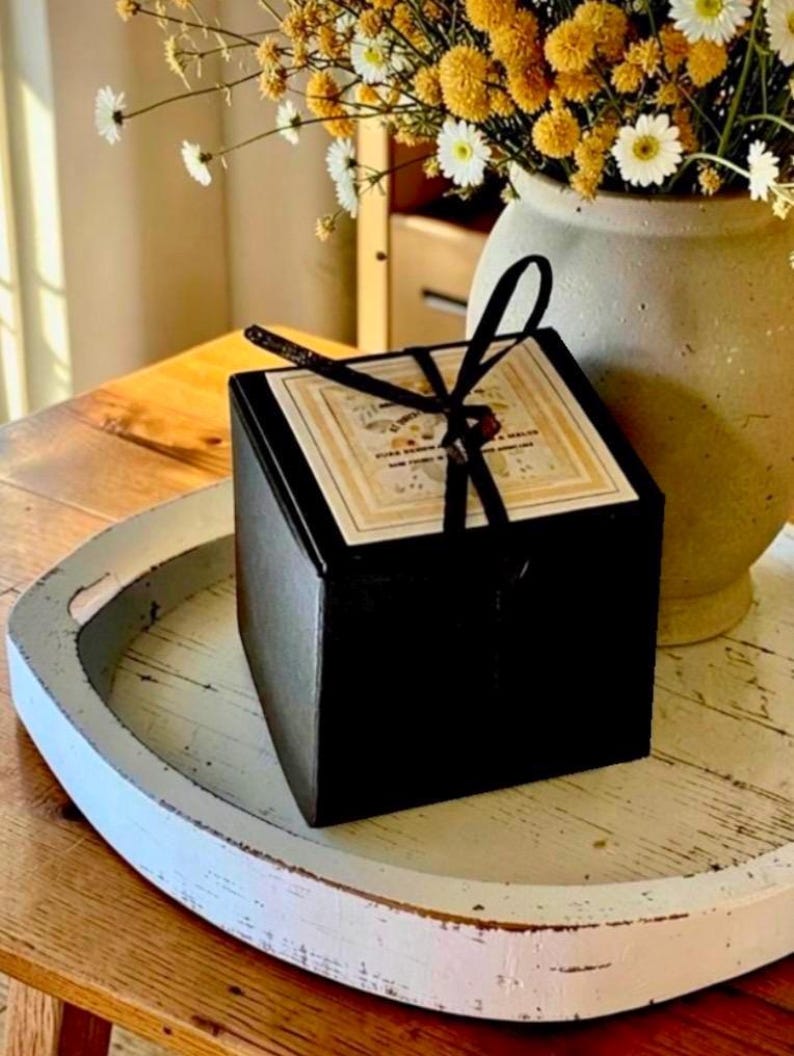 May include: A square black gift box with a black ribbon tied in a bow on top. The box features a square label with printed text and a floral design. The box is positioned on a white circular tray next to a ceramic vase filled with yellow and white flowers.