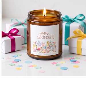 May include: A lit candle in an amber glass jar with a white label featuring the text Happy Birthday and a watercolor floral illustration. The label includes the text At Birch Hill Candle Co. The candle is surrounded by small gift boxes with colorful ribbons.
