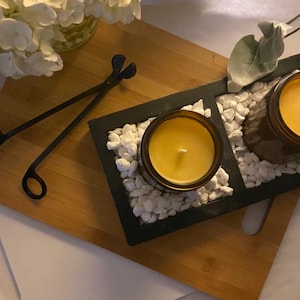 May include: Two amber glass jars filled with yellow wax candles sit inside a black rectangular tray filled with white decorative stones. A pair of black metal wick trimmers lies on a wooden board next to the tray, with white flowers nearby.