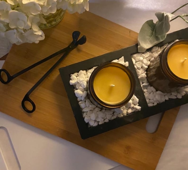 May include: Two amber glass jars filled with yellow wax candles sit inside a black rectangular tray filled with white decorative stones. A pair of black metal wick trimmers lies on a wooden board next to the tray, with white flowers nearby.