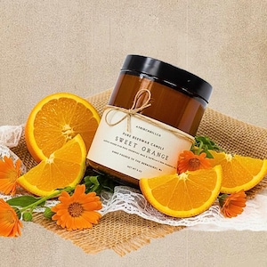 May include: A brown glass jar candle with a black lid and a cream label reading ATBIRCHHILLCO PURE BEESWAX CANDLE SWEET ORANGE NET WT 8 OZ. The candle is surrounded by fresh orange slices and bright orange calendula flowers on burlap fabric.