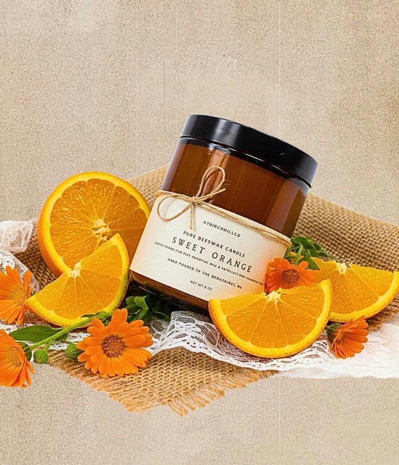 May include: A brown glass jar candle with a black lid and a cream label reading ATBIRCHHILLCO PURE BEESWAX CANDLE SWEET ORANGE NET WT 8 OZ. The candle is surrounded by fresh orange slices and bright orange calendula flowers on burlap fabric.
