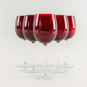 Puede incluir: Seis copas de vino tinto con tallos y bases transparentes dispuestas en semicírculo. Las copas tienen un color rojo intenso y una forma clásica. El fondo es blanco.