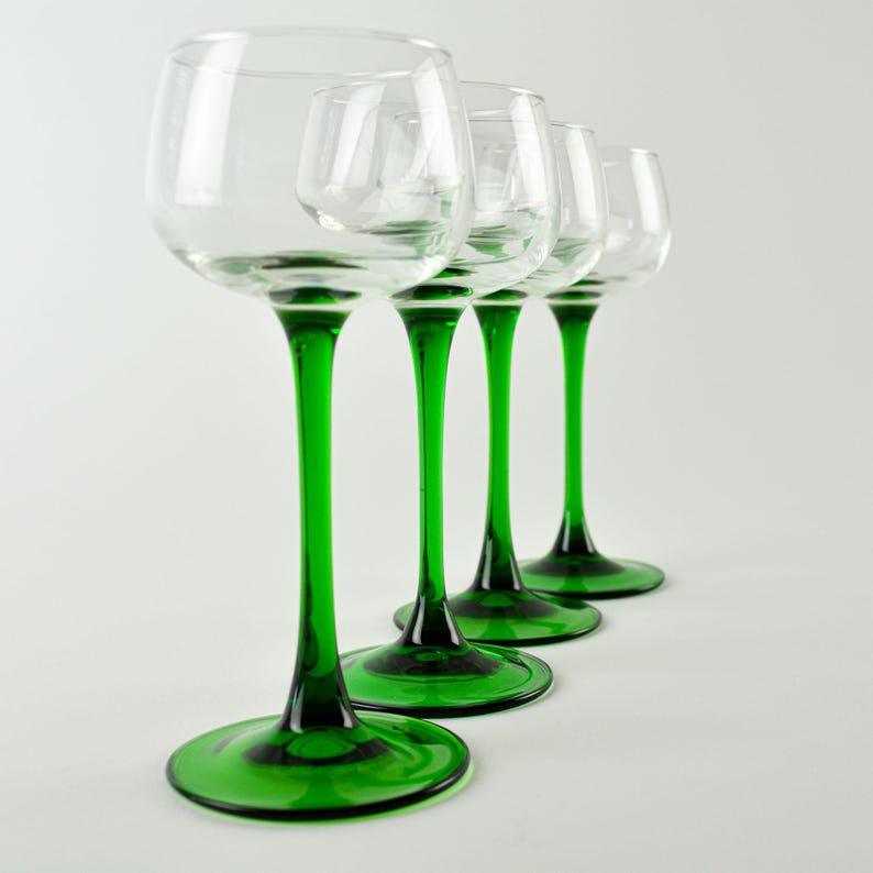 May include: Four clear wine glasses with green stems and bases are arranged in a row. The glasses have a rounded bowl shape and a long, slender stem. The green color of the stems and bases provides a striking contrast to the clear glass.