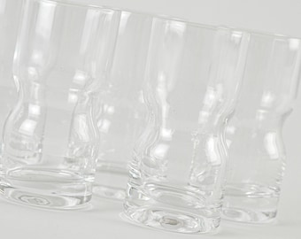 May include: A collection of clear, stemless drinking glasses. The glasses have a unique hourglass shape with a wide base and a slightly flared rim. The transparent design allows for easy viewing of the contents. The glasses are arranged on a white surface.