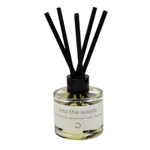 Op de afbeelding: Een glazen diffuser met zwarte rietjes en een label met de tekst "into the woods lily of the valley - sandalwood - amber - bergamot".