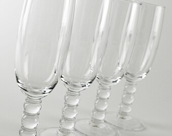 May include: Four clear champagne flutes with a rounded stem design. The glasses are arranged in a diagonal line, showcasing their elegant shape and clarity. The stems feature a series of small, rounded beads.