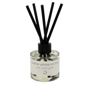 Op de afbeelding: Een diffuser van helder glas met zwarte rietjes en een wit label met de tekst "subtle white orchid vanilla - orchid - magnolia".