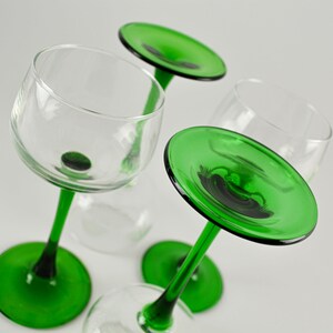 May include: Four clear wine glasses with green stems and bases are arranged on a white surface. The glasses are tilted at various angles, showcasing their design. The green color adds a pop of vibrancy.