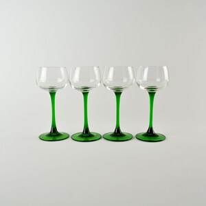May include: Four clear glass cordial glasses with green stems and bases. The glasses have a rounded bowl shape and are evenly spaced against a white background. The green color is a vibrant, rich hue.