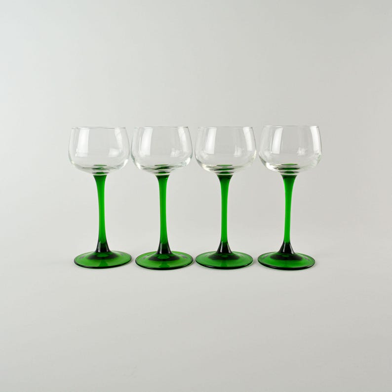 May include: Four clear glass cordial glasses with green stems and bases. The glasses have a rounded bowl shape and are evenly spaced against a white background. The green color is a vibrant, rich hue.