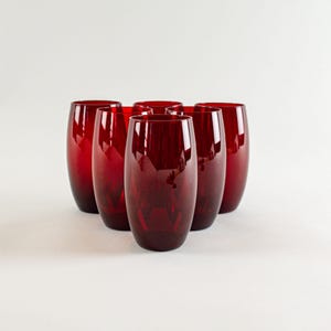 Op de afbeelding: Zes robijnrode wijnglazen zonder voet. De glazen zijn hoog, ovaal van vorm en gemaakt van een glanzend, transparant materiaal. De glazen zijn in een halve cirkel gerangschikt, waarbij het licht op hun oppervlakken weerkaatst. Het glaswerk is geschikt voor het serveren van dranken.