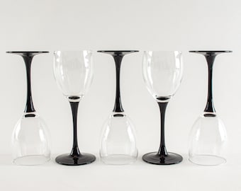 May include: Five wine glasses with clear bowls and black stems and bases are arranged in a row. The glasses are positioned with their bowls facing down. The background is plain white.