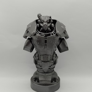 Pode incluir: Um busto de armadura de potência da série de videogames Fallout, impresso em 3D, de cor cinza metálico. O busto é detalhado com um capacete, uma placa de peito e braços.