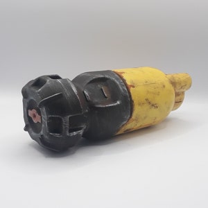 May include: A weathered, cylindrical object with a black, geometric end and a yellow body. The object has a textured appearance, suggesting it may be made of metal or a similar material. The design is reminiscent of industrial or steampunk aesthetics.