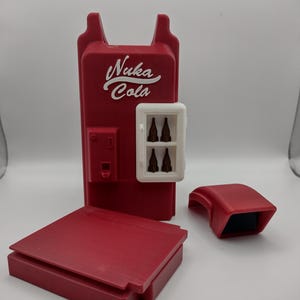 May include: A red Nuka Cola vending machine model with a white logo and a white display window containing four brown bottles. The set includes a separate red rectangular base and a detached red curved top piece, all featuring a matte finish.