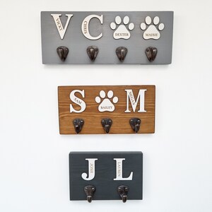 May include: Three wooden wall-mounted coat racks in gray, brown, and dark gray. Each rack has metal hooks and personalized white lettering. The top rack has the names Vicky, Chris, Dexter, and Maisie. The middle rack has Sam, Bailey, and Megan. The bottom rack has Josh and Lauren.