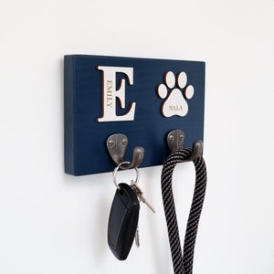 May include: A navy blue wooden key and leash holder with two metal hooks. The holder features a large letter "E" with the name "EMILY" and a paw print with the name "NALA". Keys and a dog leash hang from the hooks.
