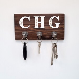 May include: A dark brown wooden key holder with three metal hooks. The key holder has the letters "CHG" in white, with the words "CAR", "HOUSE", and "GARAGE" below each letter. Keys hang from each hook.