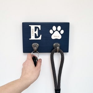 May include: A navy blue wooden key holder with two metal hooks. The key holder has a white paw print and the name "Nala". The letter "E" and the name "Emily" are also displayed. A leash hangs from one hook, and keys hang from the other.