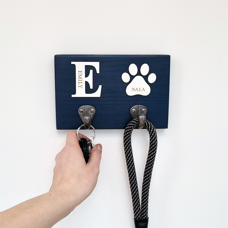 May include: A navy blue wooden key holder with two metal hooks. The key holder has a white paw print and the name "Nala". The letter "E" and the name "Emily" are also displayed. A leash hangs from one hook, and keys hang from the other.