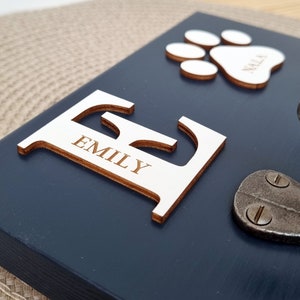 May include: A navy blue wooden plaque with a large, light-colored wooden letter "E" with the name "EMILY" engraved on it. A paw print and the name "NALA" are also present. A metal clasp is visible.