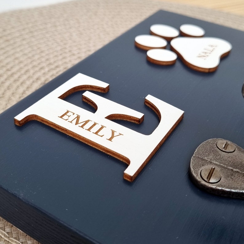 May include: A navy blue wooden plaque with a large, light-colored wooden letter "E" with the name "EMILY" engraved on it. A paw print and the name "NALA" are also present. A metal clasp is visible.