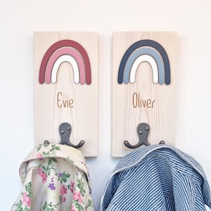 May include: Two wooden coat racks, each with a rainbow design. One features a pink, white, and red rainbow and the name "Evie". The other has a blue, white, and grey rainbow and the name "Oliver". Both have metal hooks for hanging coats.