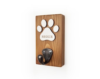 May include: A rectangular wooden wall hook featuring a medium brown stained finish. Mounted on the wood is a white paw print cutout with the name REGGIE printed in brown letters. Below the paw print is a dark gray metal hook with two screws.