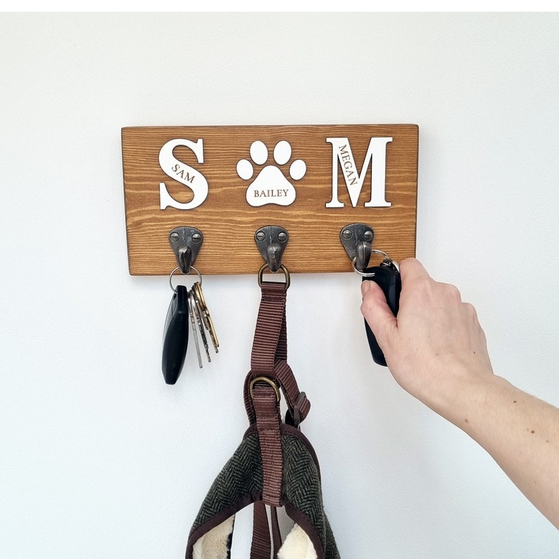 May include: A wooden key holder with three metal hooks. The key holder has the letters "S" and "M" with a paw print in the middle. The name "Bailey" is inside the paw print. Keys and a dog harness hang from the hooks.
