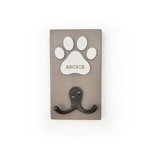 May include: A rectangular grey wooden wall hook featuring a white paw print cutout centred above a dark metal double hook. The name ARCHIE is printed in brown capital letters across the centre of the white paw print design.