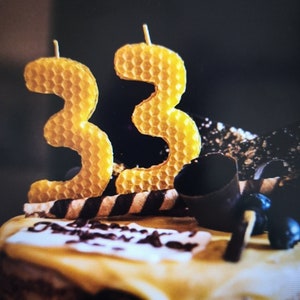 Puede incluir: Un pastel de chocolate con glaseado blanco y velas amarillas con forma de número 33 con patrón de panal en la parte superior. El pastel está decorado con rizos de chocolate y caramelos rayados en blanco y negro.