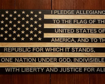 May include: A wooden sign with the Pledge of Allegiance to the United States of America. The sign is painted black with white text and features a stylized American flag with white stars on a black background.