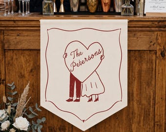 May include: A cream-colored fabric banner featuring a dark red line drawing of two figures holding a large heart with the text The Petersons written inside. The banner has a decorative border and is suspended from a wooden bar on a wooden bar counter.