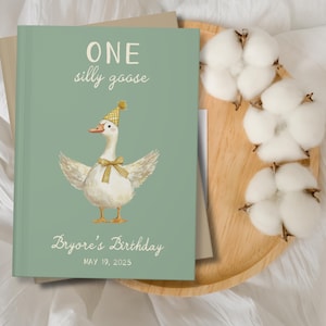 Puede incluir: Un libro de cumplea&ntilde;os verde claro con el texto "ONE silly goose" y "Bryore's Birthday MAY 19, 2025". La portada presenta una ilustraci&oacute;n en acuarela de un ganso con gorro de fiesta y pajarita. El libro est&aacute; sobre una bandeja de madera con algod&oacute;n.